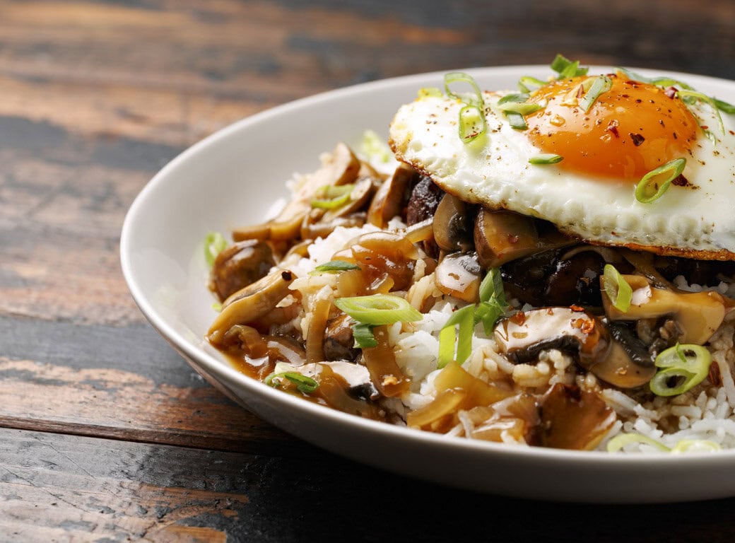 A bowl of rice topped with a fried egg, sautéed mushrooms, ground meat, brown gravy, and sliced green onions on a wooden surface—perfect comfort food after exploring things to do in Juneau.
