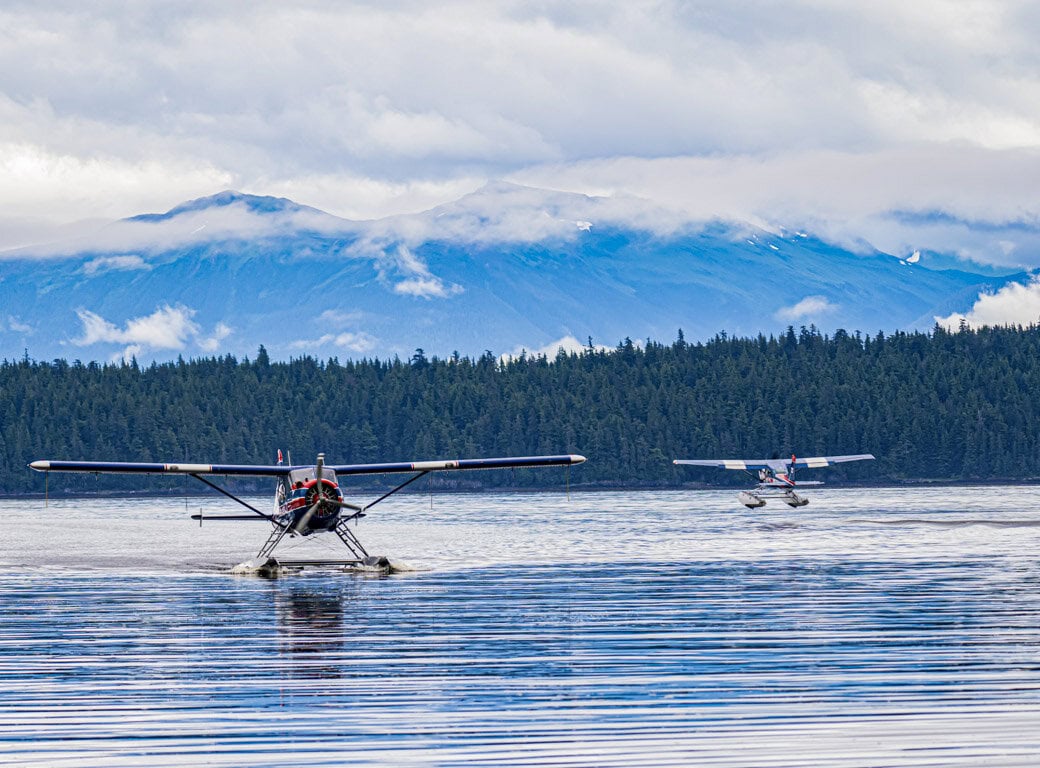 Two seaplanes are on a lake near Juneau, one taxiing and one approaching, with a forest and mountains in the background under a cloudy sky—a scene that highlights one of the top attractions and things to do in the area.