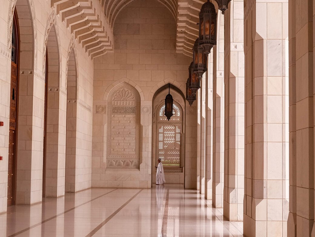 A person in white stands in the corridor of a grand mosque with tall arches, columns, and intricate wall designs, echoing the serene beauty of Oman's mountains.