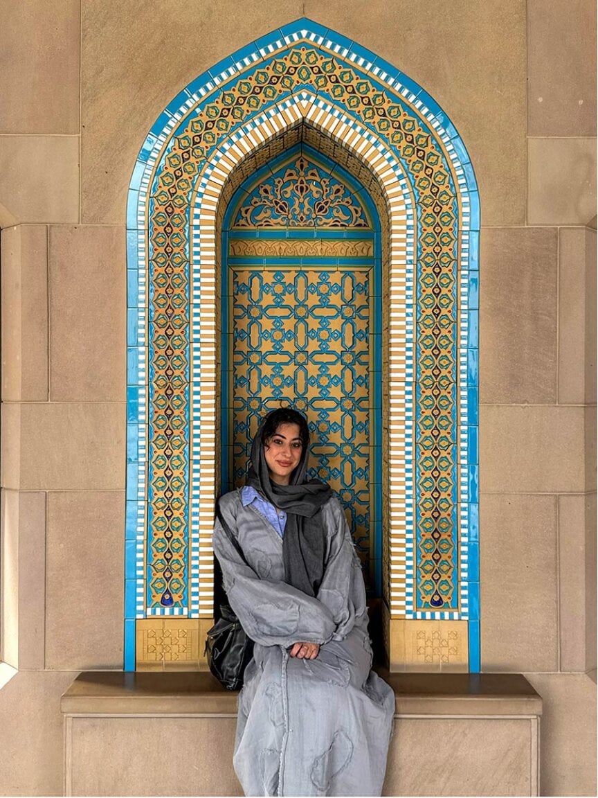 A woman wearing a gray abaya and headscarf sits in front of a decorated arched niche with blue and gold geometric patterns, reminiscent of designs found near Oman's desert and rugged mountains.
