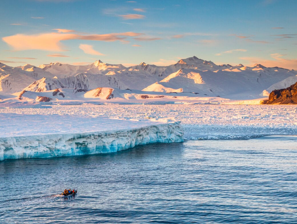 A small boat with people approaches a large ice shelf on calm water during an Antarctica tour, with snow-covered mountains and a colorful sky in the background.