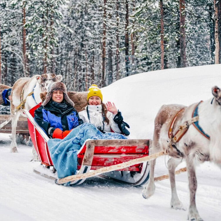 Two people sit in a red sleigh pulled by reindeer through a snowy forest, bundled in winter clothes and blankets; one person waves at the camera, capturing one of winter’s must-see moments.