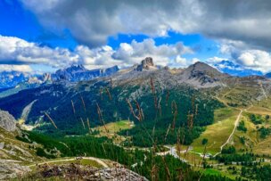 Mountain landscape with rugged peaks, green valleys, forested areas, and winding roads under a partly cloudy sky. Perfect for hiking in the Dolomites in September, with tall grass swaying in the foreground.