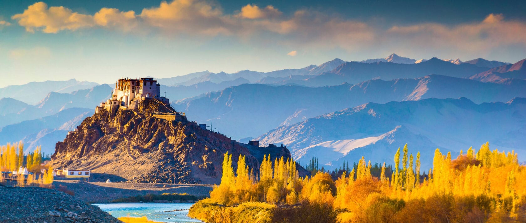 A monastery sits atop a rocky hill, surrounded by autumn trees and a river, with layers of blue mountains in the background under a partly cloudy sky.