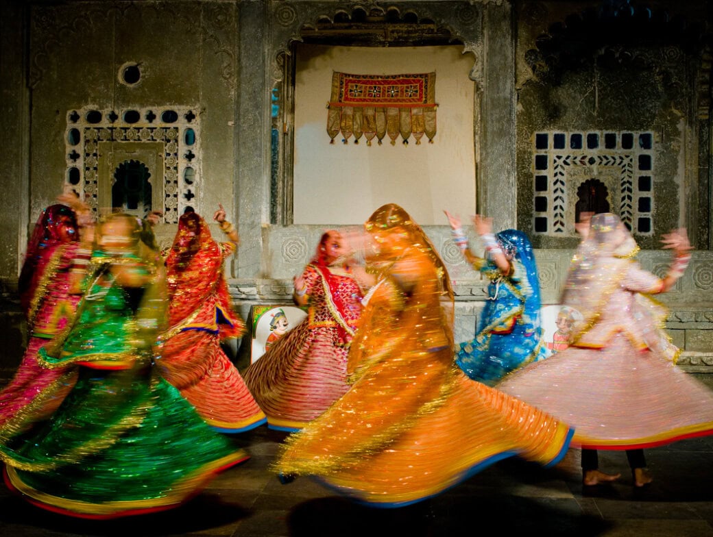 A group of women in colorful traditional attire perform a spinning dance in front of ornately decorated walls with arched windows—moments like these are not to miss in 2026.