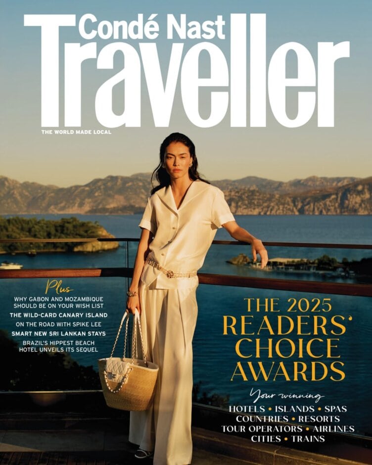 A woman in a cream outfit stands by a wooden railing with a scenic lake and mountains in the background on the cover of Condé Nast Traveller, celebrating the 2025 Readers' Choice Awards and top tour operators.