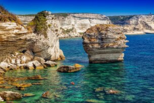 Large limestone cliffs and rock formations rise from clear turquoise water along a rugged coastline under a bright blue sky—one of the Top Ten Destinations to explore for Travel 2026.