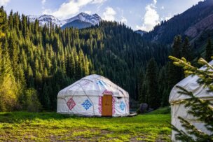 A traditional yurt with decorative patterns stands on grass in a forested mountain landscape, perfect for Travel 2026, with pine trees and snow-capped peaks offering one of the Top Ten Destinations for nature lovers.
