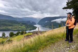 A person in hiking gear stands on a grassy hillside overlooking a fjord, with mountains and cloudy skies in the background, perhaps pausing to enjoy some traditional Norwegian foods along the way.