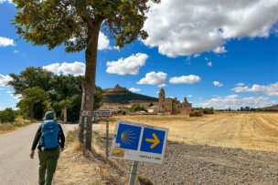 A person with a backpack walks along a rural road toward a village, passing an el camino tours sign under a tree with a blue sky and scattered clouds overhead.