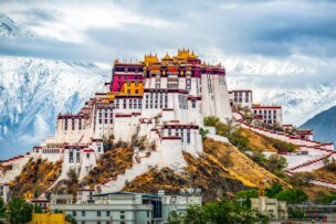 A large, multi-leveled fortress with white walls and golden roofs sits on a hilltop, framed by snow-capped mountains under a cloudy sky—a striking landmark among the Top Ten Destinations for Travel 2026.