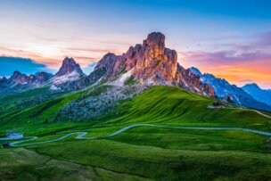 A winding road leads through green hills toward jagged mountain peaks under a colorful sunset sky—one of the best places to visit for travel 2026.
