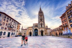 Two people with backpacks stand in a large, empty plaza before a historic stone cathedral—one of the top destinations 2026—its tall spire reaching up among old buildings under a partly cloudy sky.