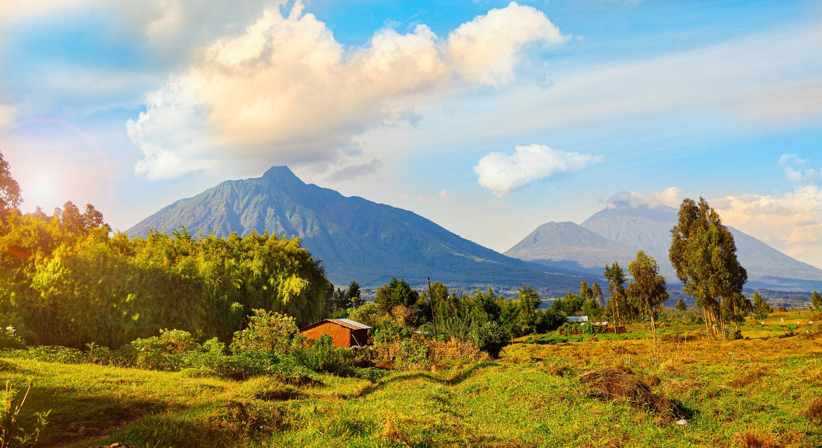 A landscape with green fields, scattered trees, small houses, and two large volcanic mountains under a partly cloudy sky, reminiscent of the serene views found in the best parks Rwanda has to offer.