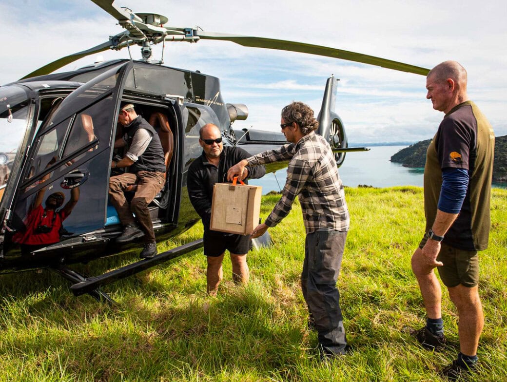 Four people from the community unload a wooden box from a black helicopter on a grassy field overlooking water and hills, supporting conservation efforts.