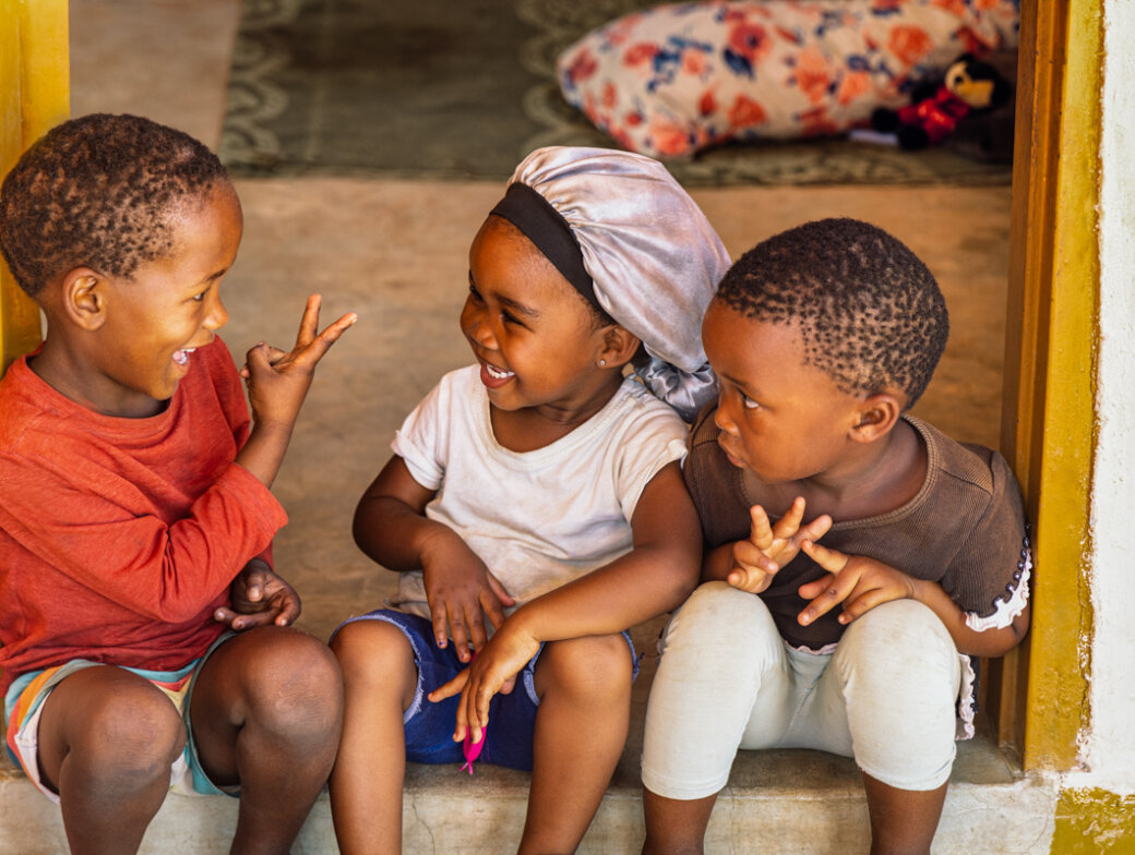 Three young children sit together in a doorway, smiling and playing hand games. A colorful pillow lies on the floor behind them, reflecting the warmth of community and shared conservation values.