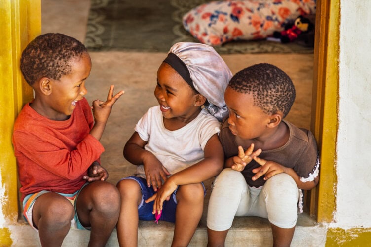 Three young children sit together in a doorway, smiling and laughing as they gesture animatedly—a joyful scene reminiscent of friendships formed on Botswana safaris.