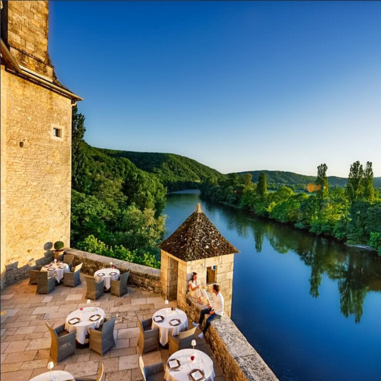 Outdoor dining tables set on a stone terrace overlook a calm river and green hills, with two people sitting near a small turret under a clear blue sky.
