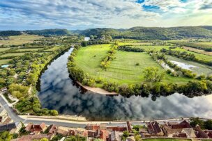 Aerial view of a river curving around a green field in Southern France, with houses and a road in the foreground and forested hills under a partly cloudy sky—perfect for a scenic stop on a cultural tour.