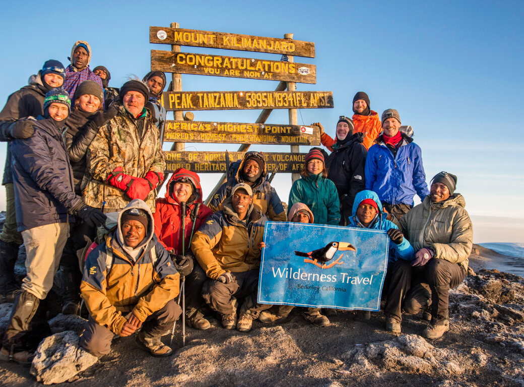 A group of people in outdoor gear pose at the Mount Kilimanjaro summit sign, celebrating community and conservation, some holding a "Wilderness Travel" banner with a clear sky and distant landscape in the background.