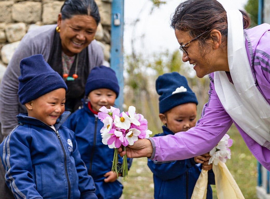 A woman in a pink jacket hands a bouquet of flowers to three smiling children in blue uniforms and hats, as another woman stands behind them outdoors, reflecting community spirit and a shared love for conservation.