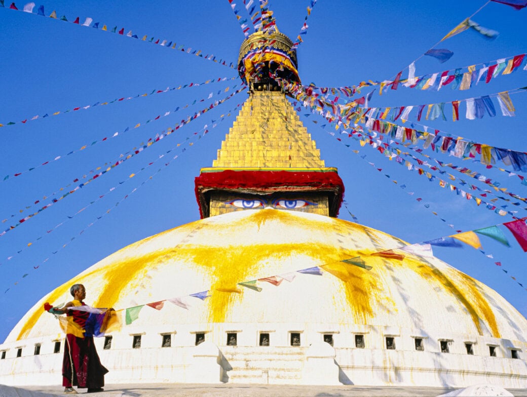 A person stands in front of the white-domed Boudhanath Stupa in Nepal, one of the top things to do in Kathmandu, decorated with prayer flags and featuring painted eyes on its central tower under a clear blue sky.