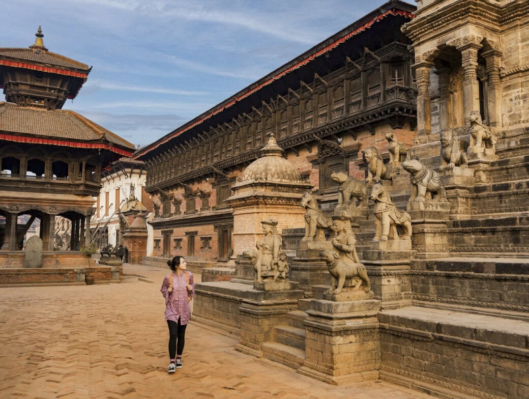 A person walks through an ancient courtyard in Kathmandu, surrounded by intricate stone statues and traditional buildings with terracotta roofs under a blue sky—a must-see among the many things to do in Kathmandu.