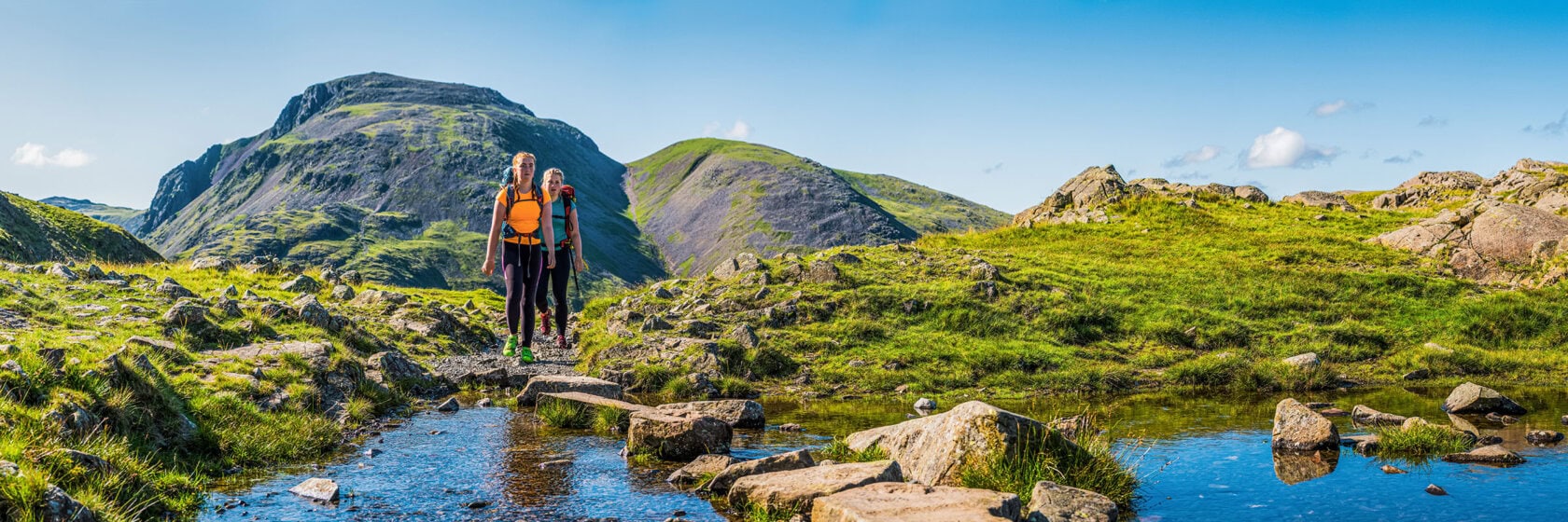 Two hikers with backpacks stand on a rocky path near a small stream, surrounded by green hills and mountains under a clear blue sky—perfect for those looking to compare England trips and soak in the scenic countryside.