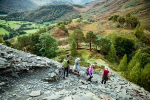 Five people walk down a rocky hillside trail surrounded by green trees and rolling hills, capturing the scenic beauty found in an England Coast to Coast Hike beginner's guide.