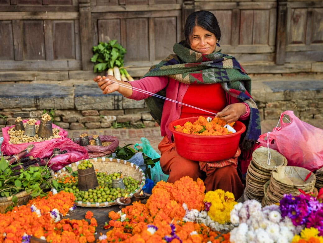 A woman in a colorful shawl sits on the ground, stringing marigold flowers, surrounded by baskets of produce at a bustling market—a vibrant scene among the many things to do in Kathmandu.