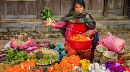 A woman in a colorful shawl sits on the ground, stringing marigold flowers, surrounded by baskets of produce at a bustling market—a vibrant scene among the many things to do in Kathmandu.