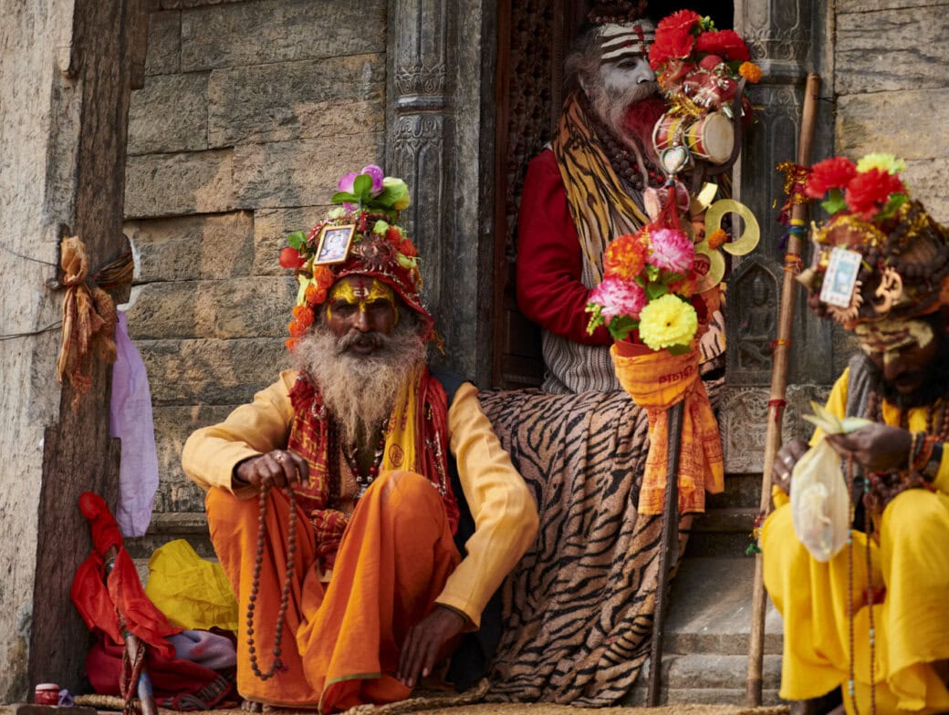 Three colorfully dressed sadhus with painted faces sit and pose near a temple entrance, adorned with garlands, flowers, and traditional accessories—a memorable sight among the top things to do in Kathmandu.
