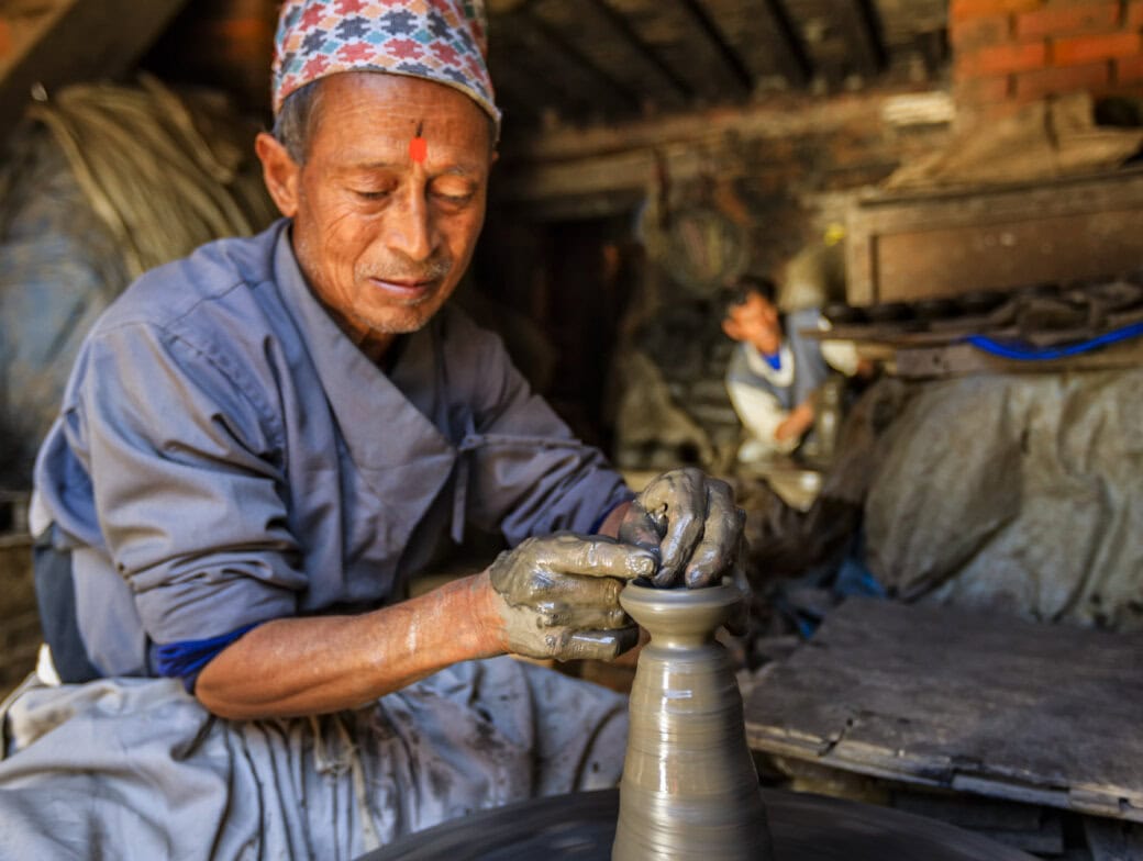 An artisan shapes clay on a pottery wheel in a rustic workshop, focusing intently on his work—one of the authentic things to do in Kathmandu—with another person visible in the blurred background.