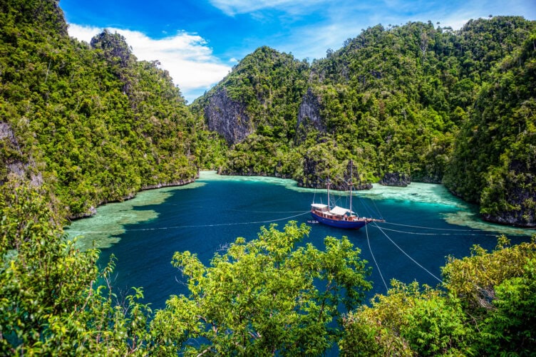 A wooden boat is anchored in clear blue water surrounded by lush, green limestone hills under a bright sky.