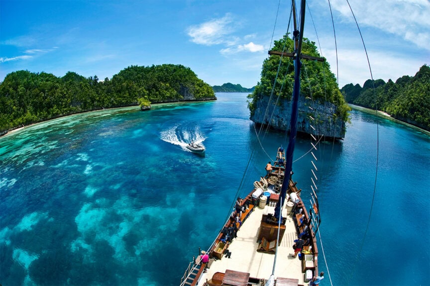 A boat cruises through clear turquoise water near green islands in Raja Ampat, viewed from the deck of a larger ship under a blue sky—an inviting scene for snorkeling or scuba diving adventures.