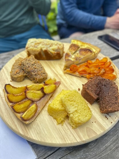 A round wooden platter with various baked goods—including quiche, carrot slices, fruit tart, cornbread, loaf cake, and brownies—awaits on an outdoor table, perfect for refueling after a hiking Tour du Mont Blanc adventure.