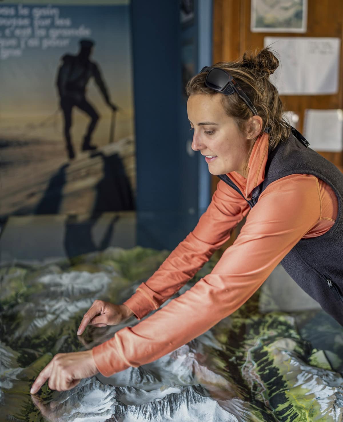 A woman in an orange jacket and sunglasses points at locations on a raised relief map of mountainous terrain indoors, possibly planning her next hiking adventure along the iconic Tour du Mont Blanc.