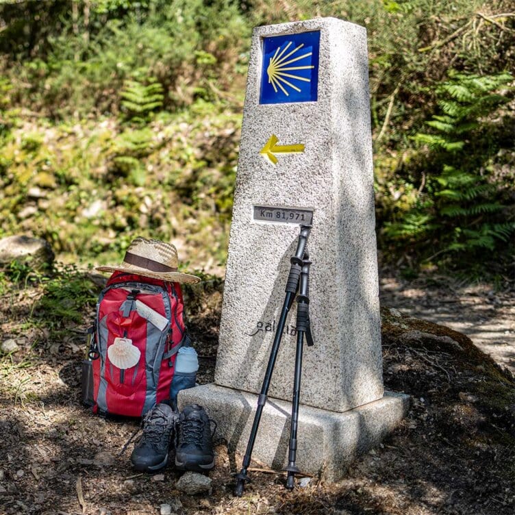 A Camino de Santiago milestone marker with a yellow arrow, backpack, hiking boots, trekking poles, and a hat placed beside it on a forest path during the Holy Year.