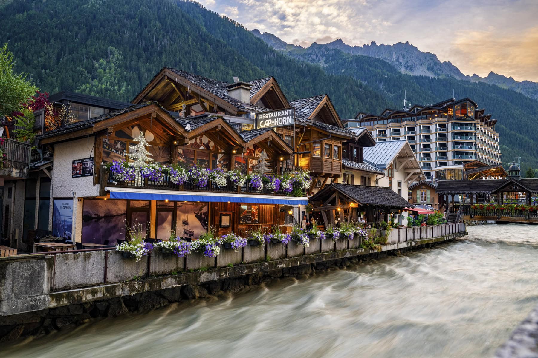 A riverside restaurant with flower boxes sits beside a flowing stream, perfect for relaxing after hiking the Tour du Mont Blanc, with mountain buildings and forested hills in the background under a cloudy sky.
