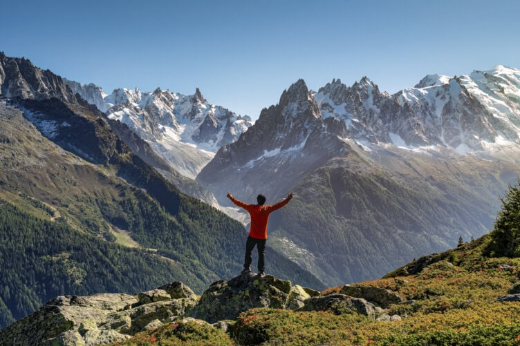 A person in a red jacket stands on a rocky ledge with arms raised, celebrating their hiking Tour du Mont Blanc, overlooking snow-capped peaks and green valleys under a clear blue sky.