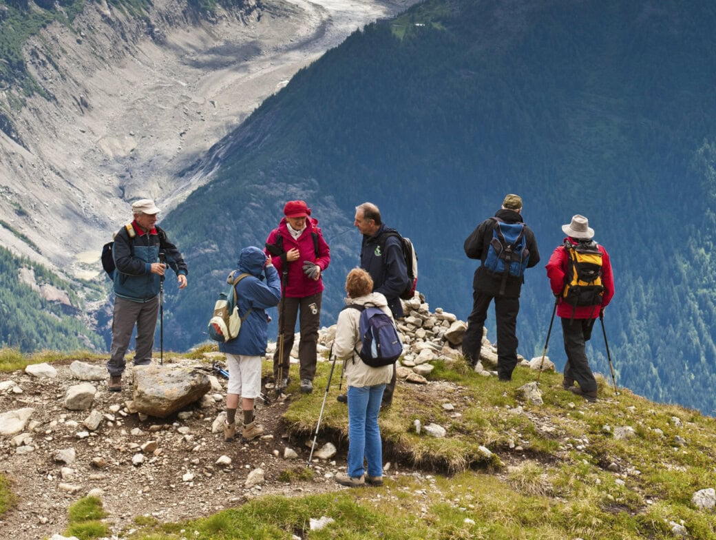A group of hikers with backpacks and walking sticks stand on a grassy mountain trail, enjoying breathtaking views along the renowned hiking Tour du Mont Blanc, overlooking a deep forested valley.