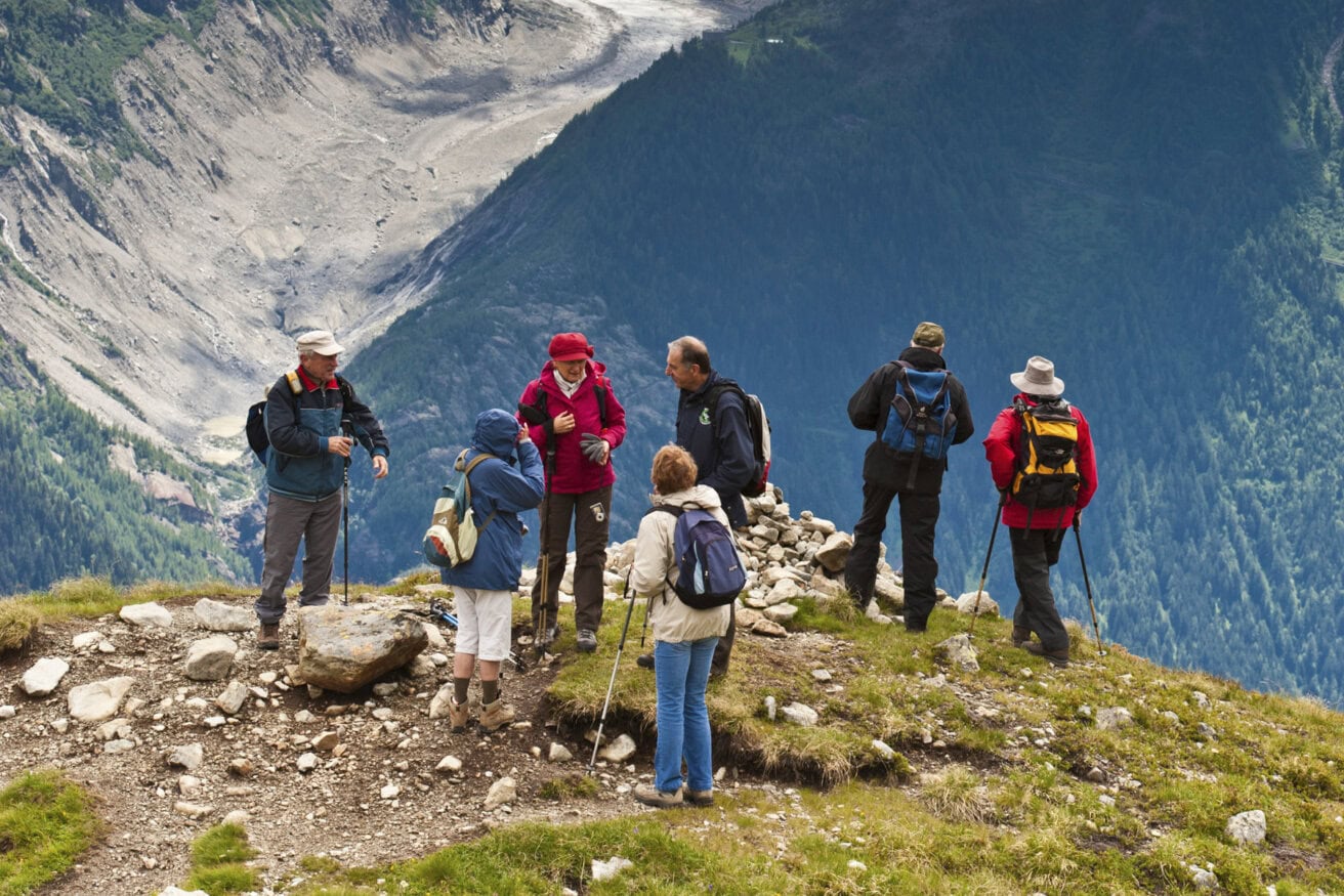 A group of hikers with backpacks and walking sticks stand on a grassy mountain trail, enjoying breathtaking views along the renowned hiking Tour du Mont Blanc, overlooking a deep forested valley.