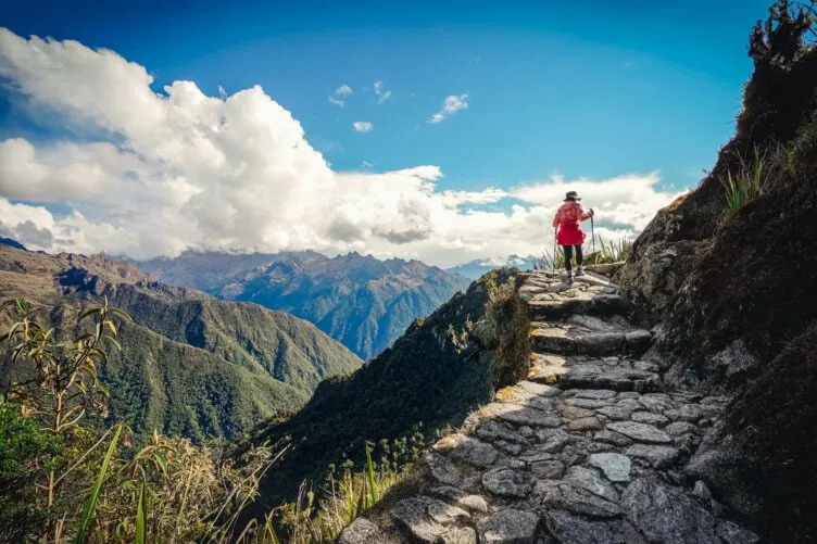 A person in a red jacket and hat walks on a stone path along a mountain ridge in Peru, with distant peaks and clouds in the background—an inspiring scene for any travel guide.