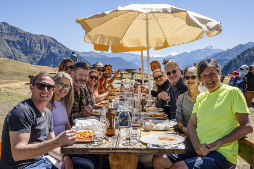 A group of people sit at a long outdoor table eating a meal together under a large umbrella, with mountains visible in the background after a day of hiking the Tour du Mont Blanc.