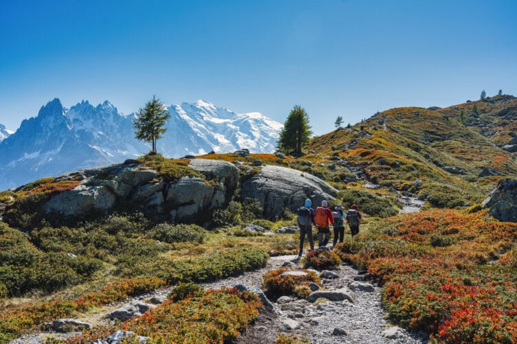 Three hikers walk along a rocky trail through an alpine landscape on the famous Tour du Mont Blanc, with snow-capped mountains and a clear blue sky in the background.