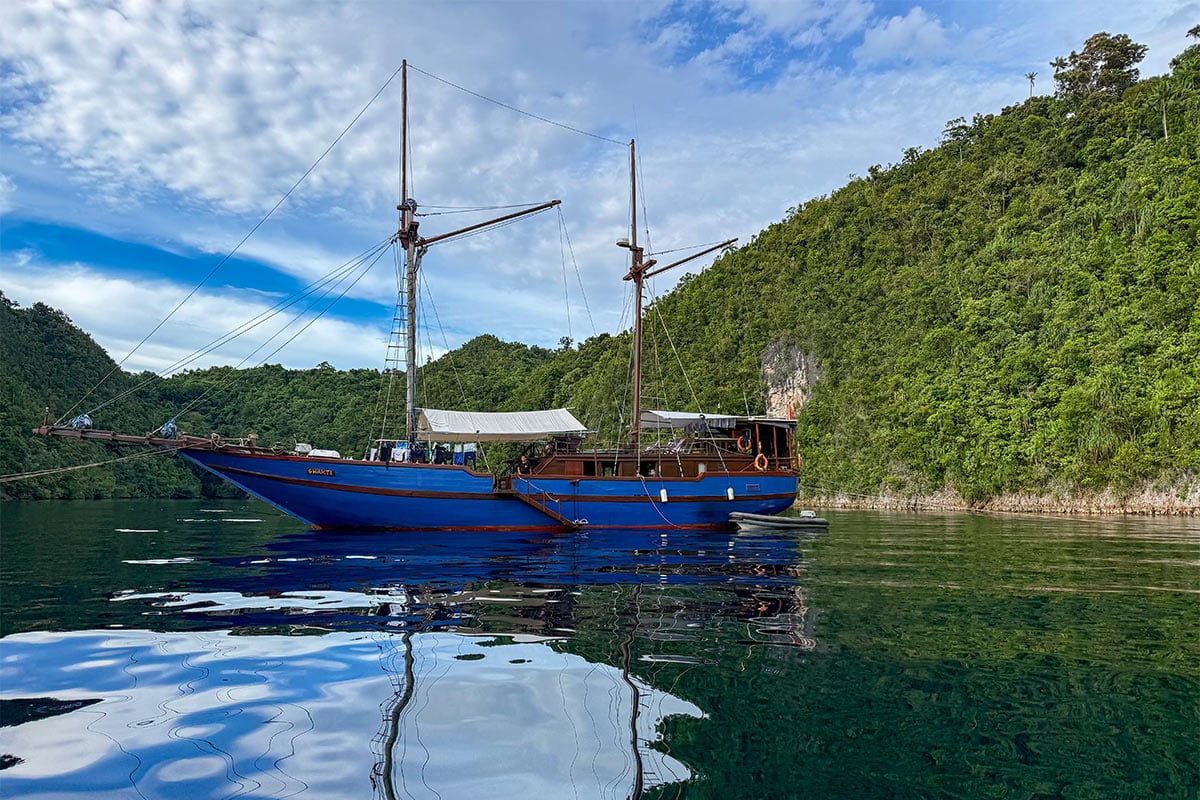 A blue wooden sailing boat with two masts floats on calm, reflective water near a lush, green forested shoreline under a partly cloudy sky—an inviting scene in Raja Ampat, renowned for its underwater paradise and exceptional snorkeling.