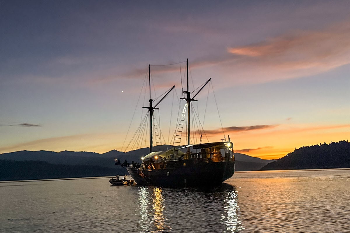 A large, two-masted ship anchored on calm water at sunset near the hills and mountains of Raja Ampat, an underwater paradise perfect for snorkeling adventures.