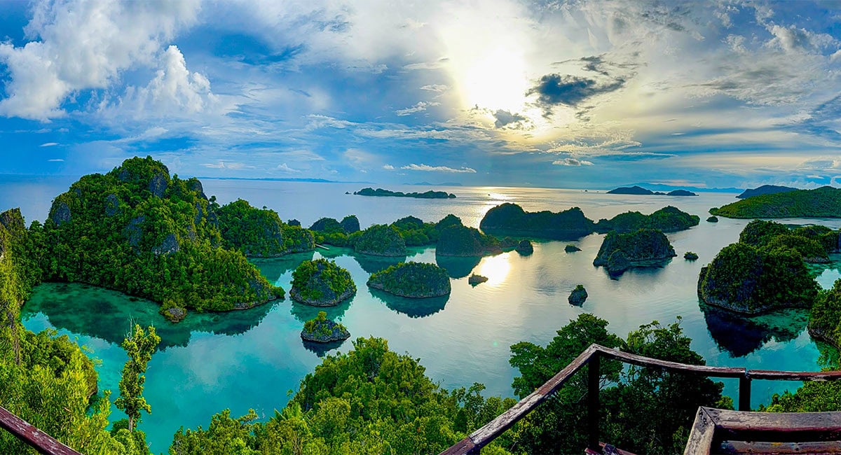 View of scattered green islands in a calm, blue sea under a partly cloudy sky with sunlight reflecting off the water—an underwater paradise in Raja Ampat, perfect for snorkeling adventures.