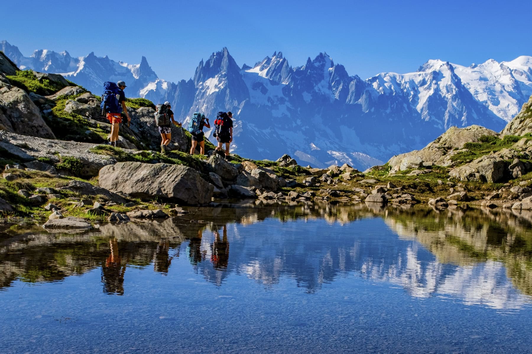 A group of hikers with backpacks stand by a mountain lake along the Tour du Mont Blanc, with snow-capped peaks and clear blue sky reflected in the water.