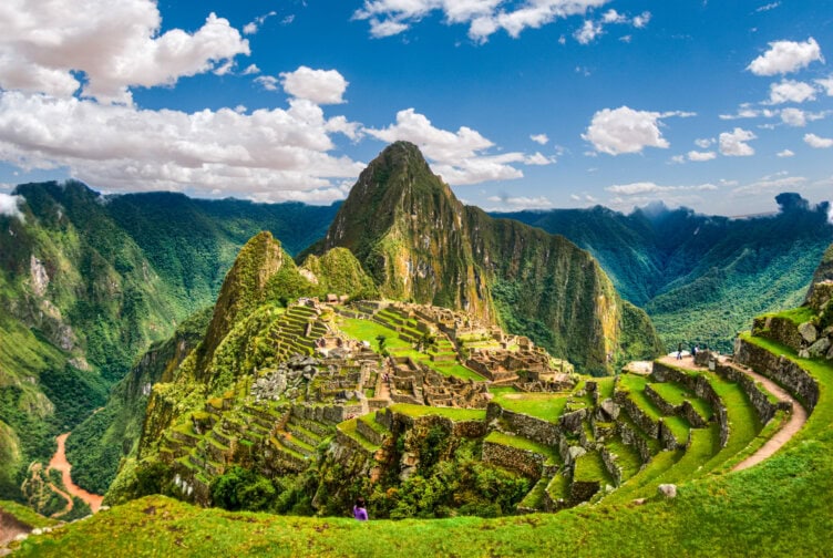 View of Machu Picchu ruins surrounded by green mountains and terraces under a partly cloudy blue sky in Peru, a must-see destination featured in every travel guide.